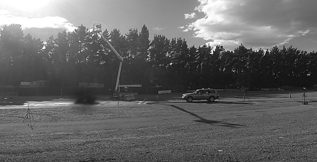 A car is parked on a gravel lot near a tall, extended construction lift, with trees and scattered clouds in the background. Some equipment is visible near the lift, and shadows stretch across the ground.
