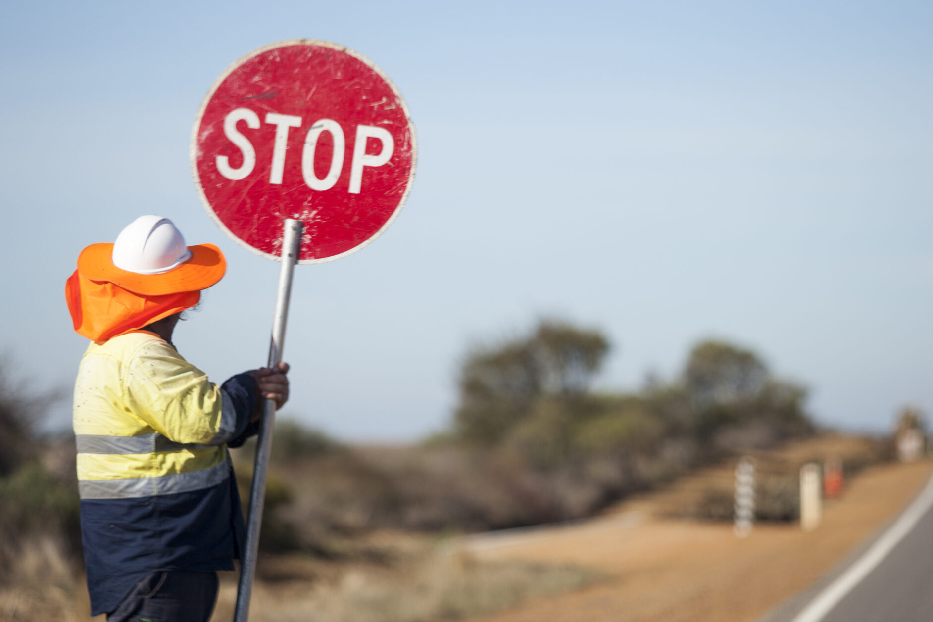 A construction worker in a high-visibility jacket and orange hat holds a red STOP sign on the side of a rural road, with blurred trees and landscape in the background.