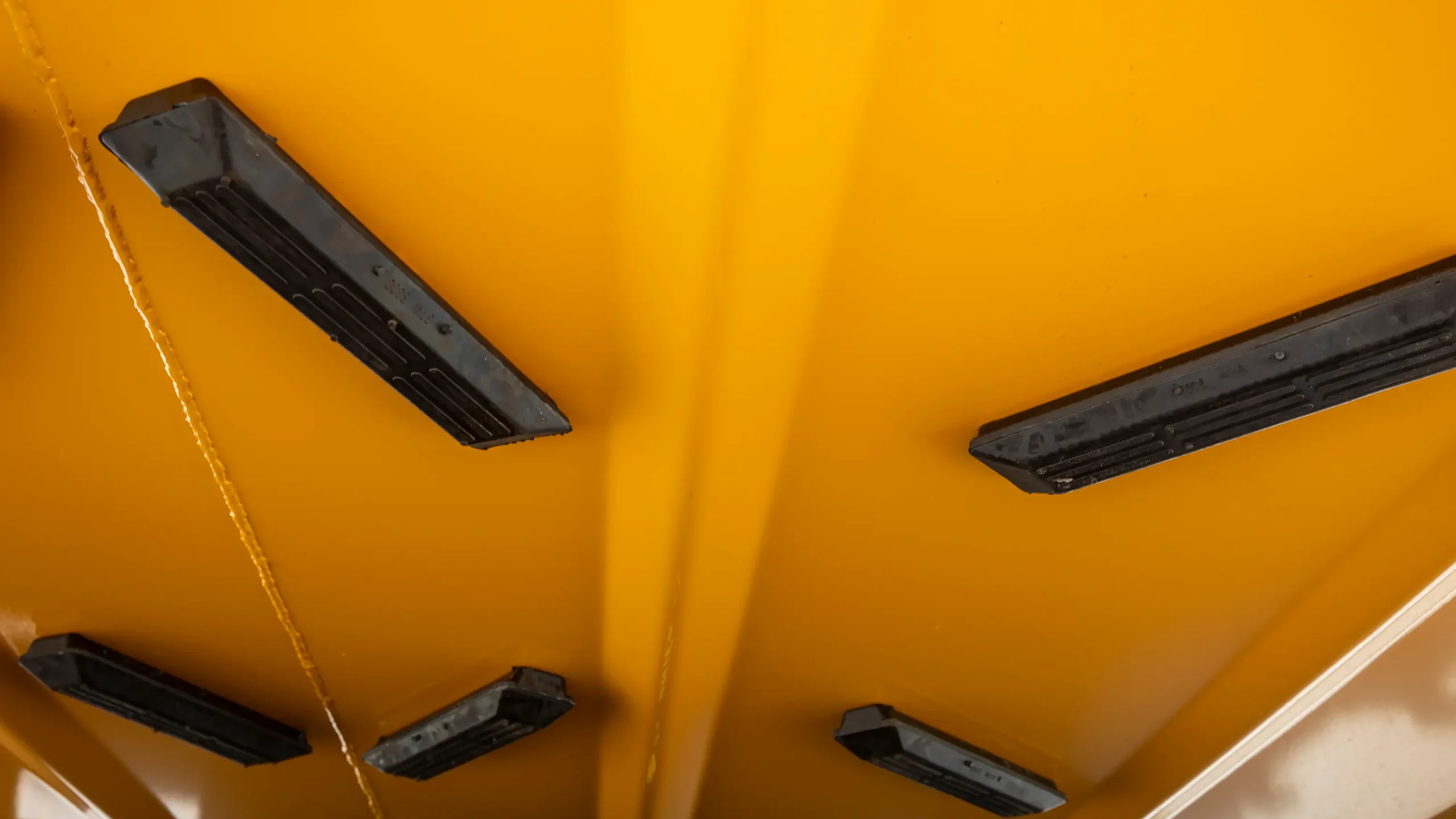 Close-up of the underside of a bright yellow structure, possibly a vehicle or industrial equipment, featuring several black rectangular rubber pads or strips evenly attached to the surface.