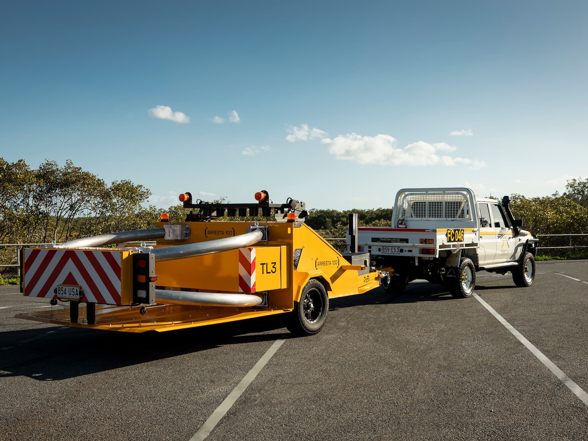 A white utility truck is towing a yellow traffic safety crash attenuator trailer in an empty parking lot, with trees and a clear sky in the background.