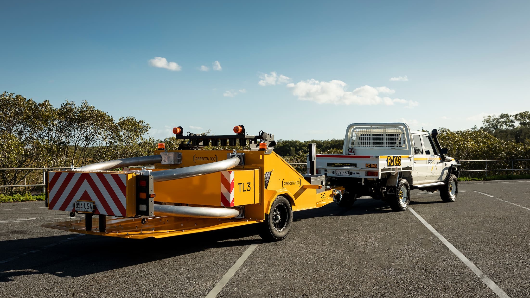 A white utility truck is towing a yellow traffic safety crash attenuator trailer in an empty parking lot, with trees and a clear sky in the background.