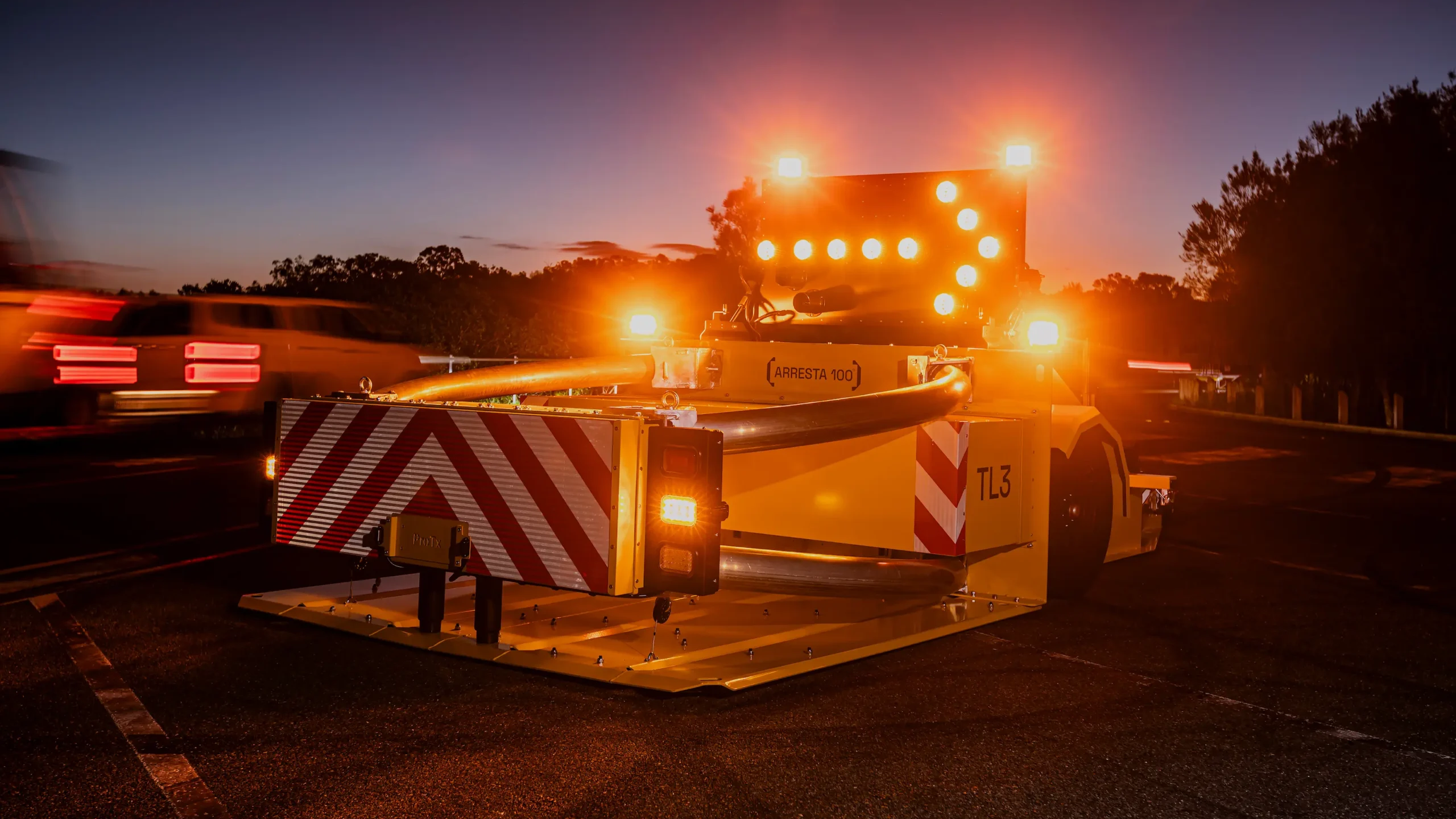A yellow crash truck with flashing orange lights and a striped crash cushion is stationed on a road at dusk, with trees and blurred car lights in the background.