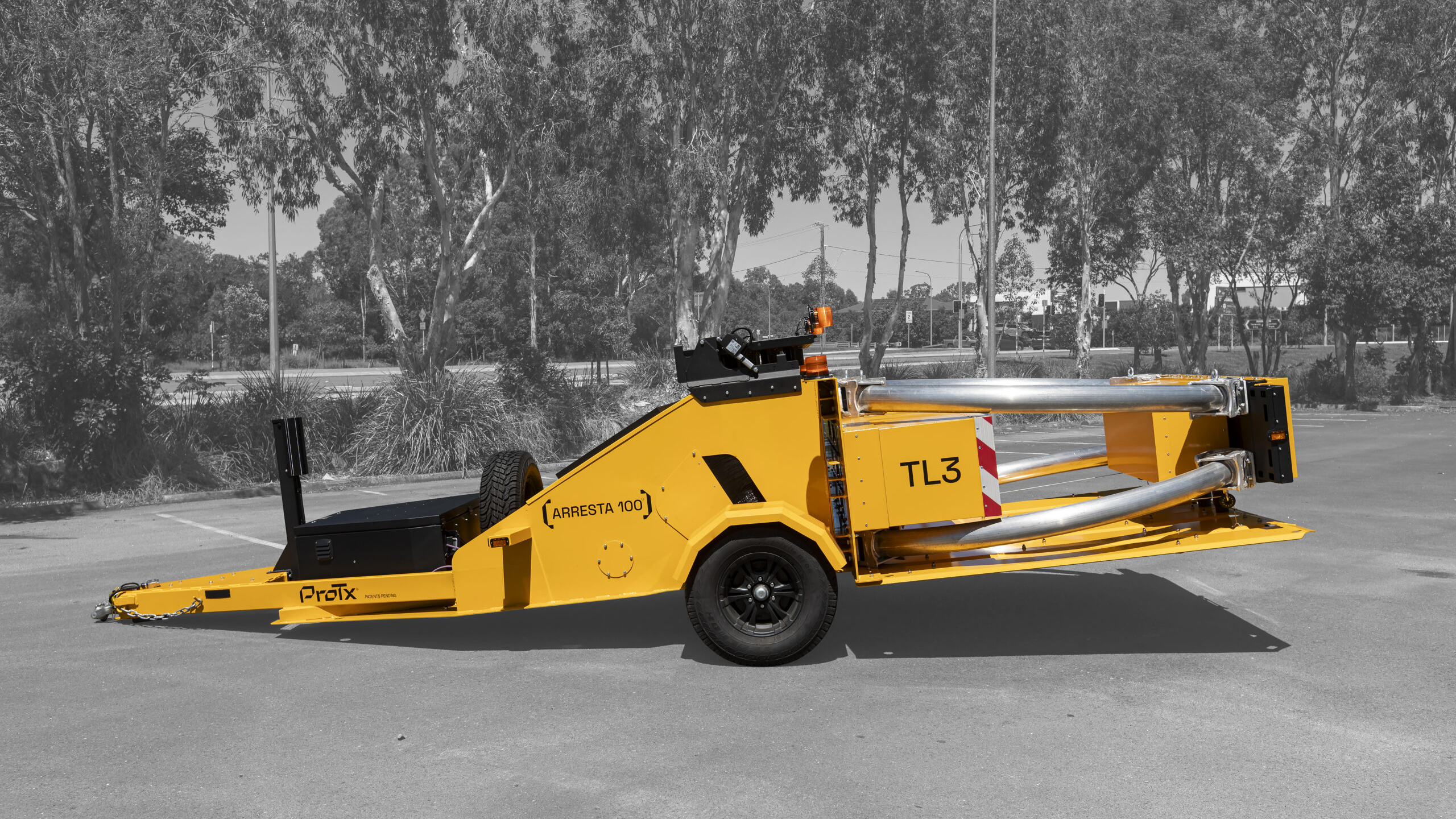 A yellow mobile crash barrier trailer marked TL3 and Arresta 100 is parked on a paved surface, with trees and light poles visible in the black-and-white background.