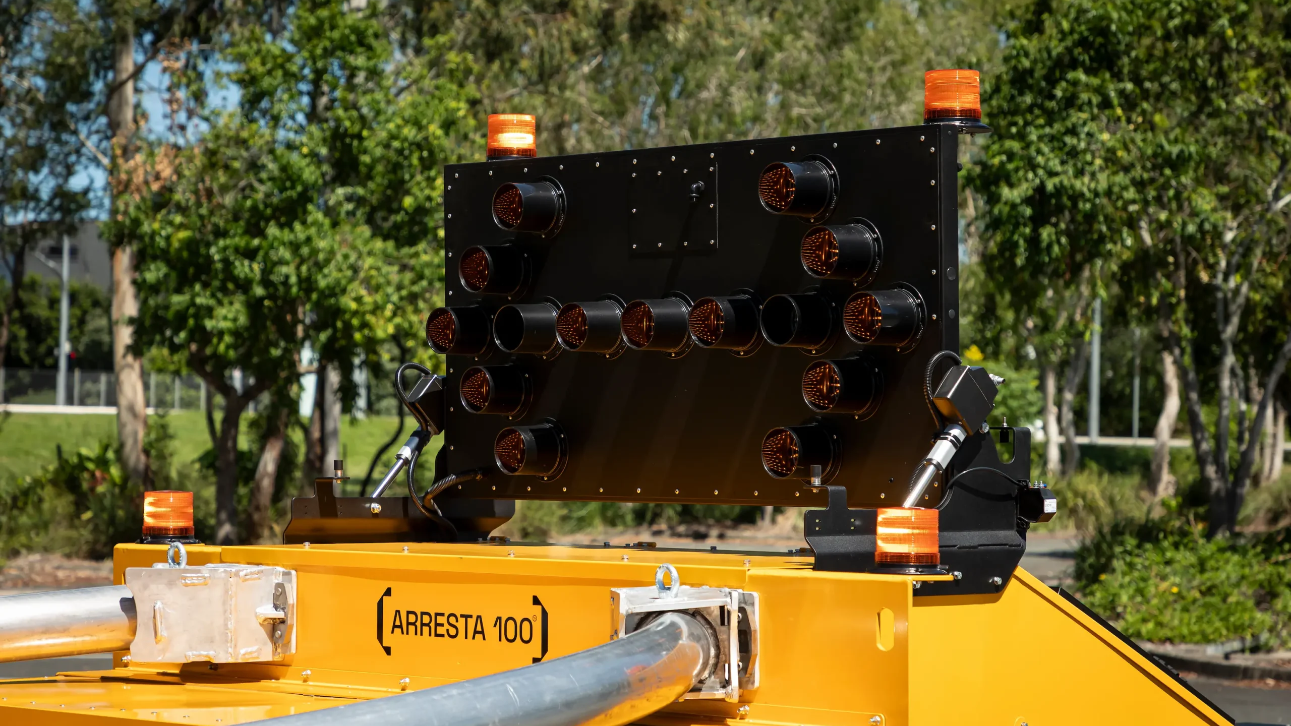 A close-up of a yellow roadside crash cushion with a mounted black electronic arrow board and orange warning lights, set outdoors with trees in the background.