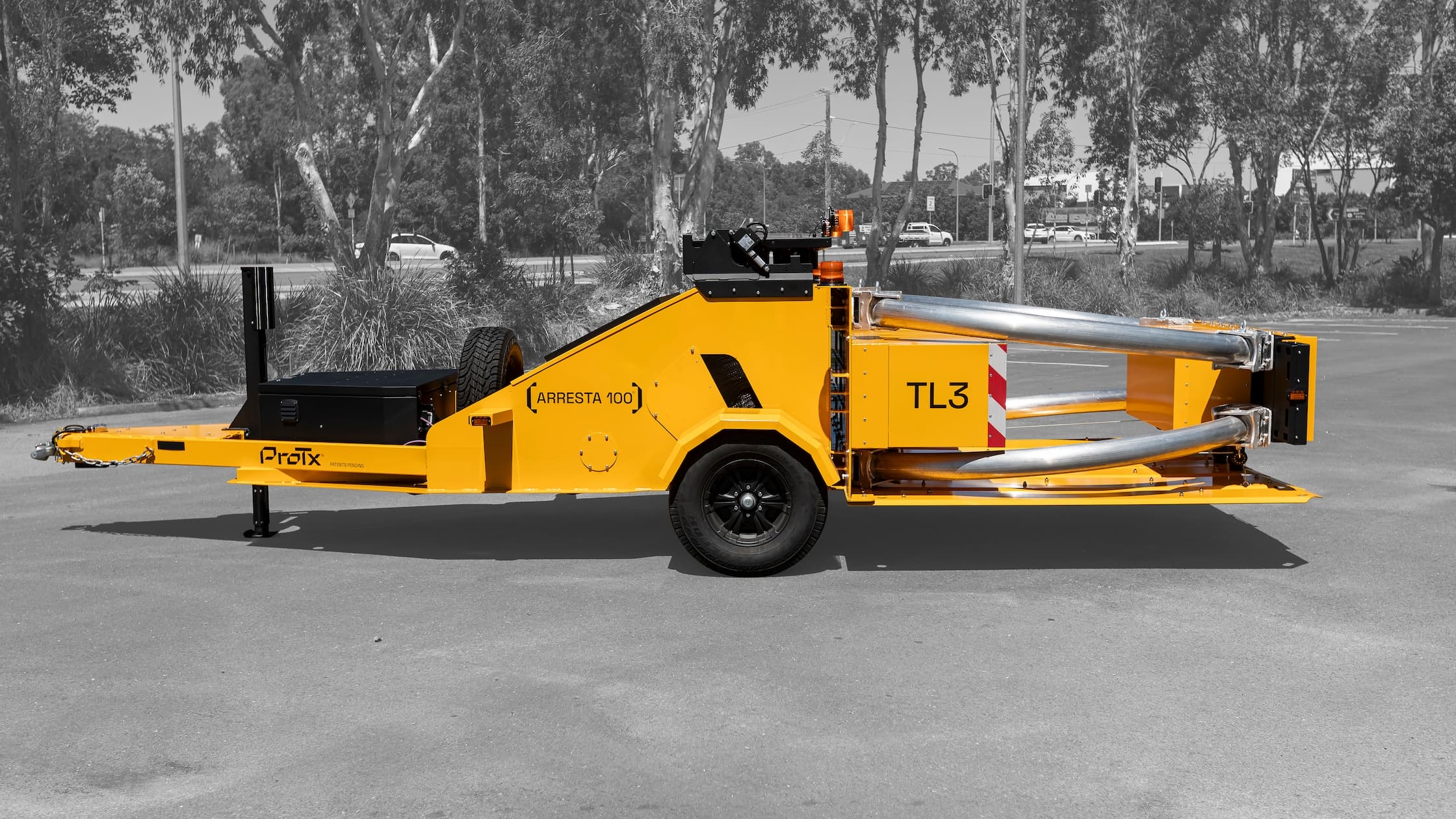 A bright yellow crash cushion trailer labeled Arresta 100 and TL3 is parked on a paved surface, with metal tubes extending from the rear. Trees and parked cars are visible in the black-and-white background.