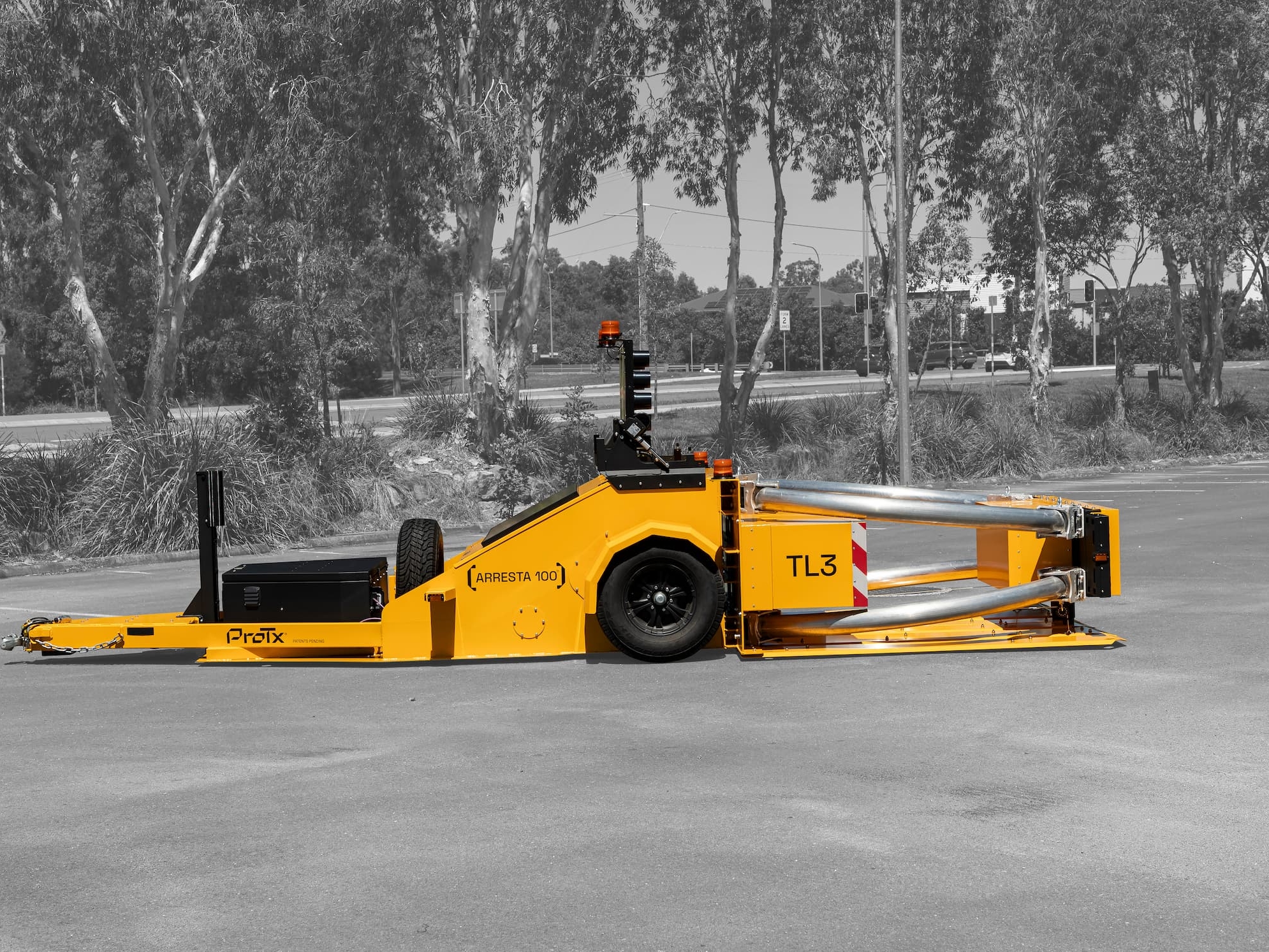 A bright yellow roadside crash attenuator trailer, labeled TL3 and ProTix, is parked on asphalt with trees and bushes in the background. The trailer is shown in a side view outdoors.