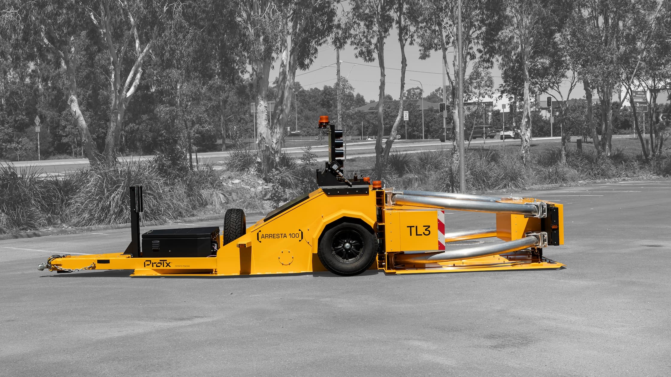 A bright yellow roadside crash attenuator trailer, labeled TL3 and ProTix, is parked on asphalt with trees and bushes in the background. The trailer is shown in a side view outdoors.