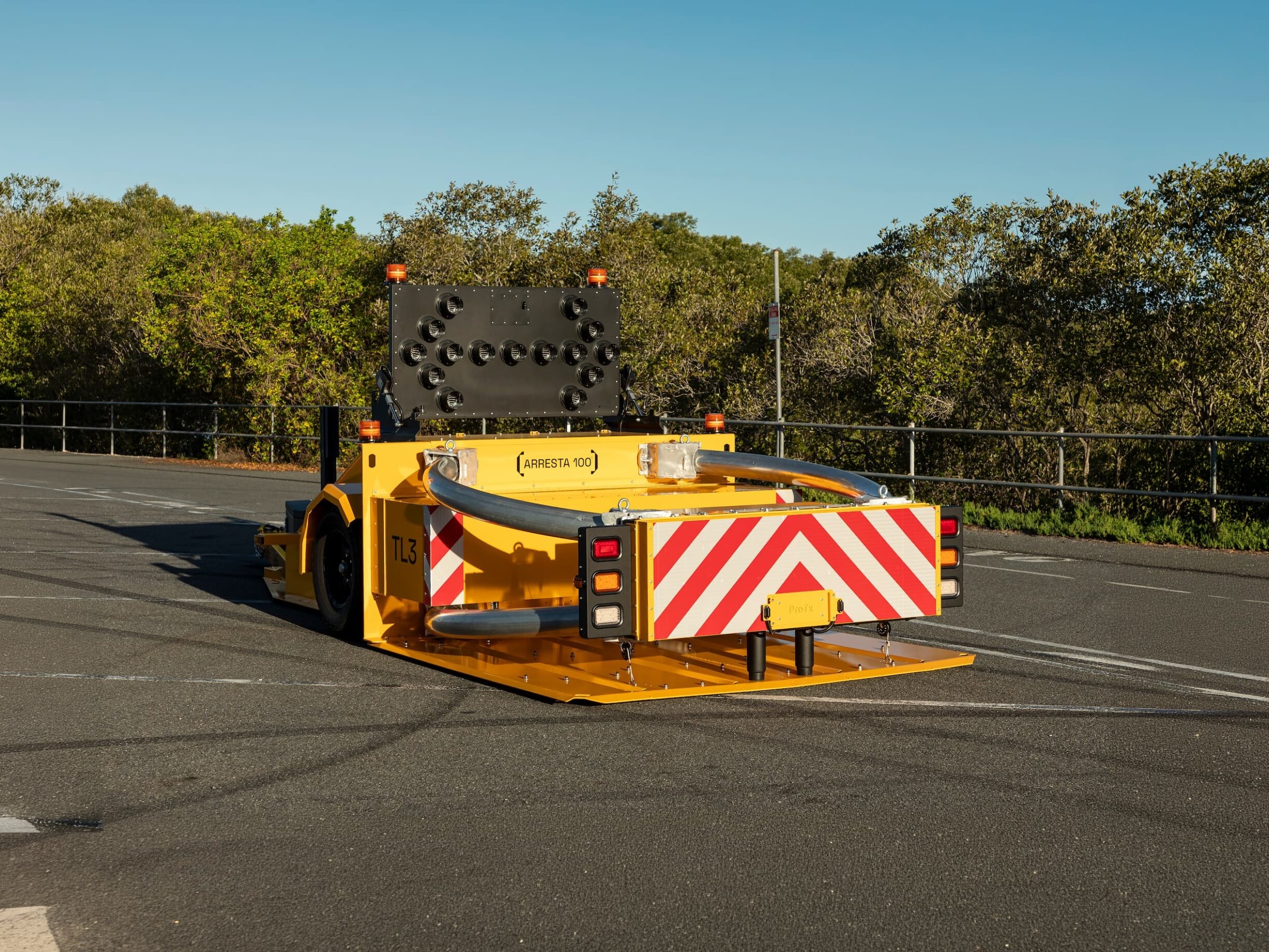 A yellow crash cushion vehicle with red and white striped warning markings is parked on asphalt near trees and a black metal fence under a clear blue sky.