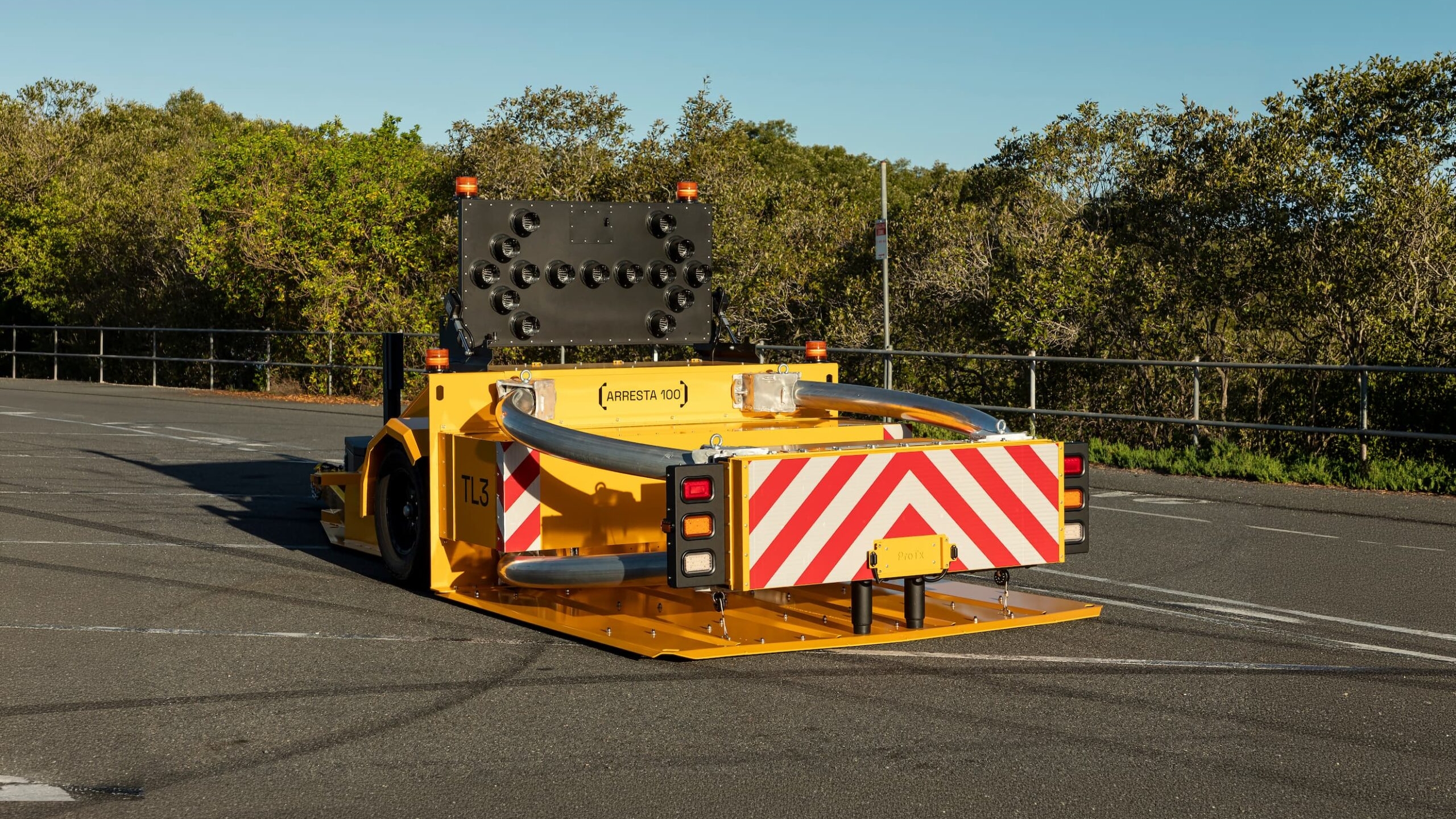A yellow crash cushion vehicle with red and white striped warning markings is parked on asphalt near trees and a black metal fence under a clear blue sky.