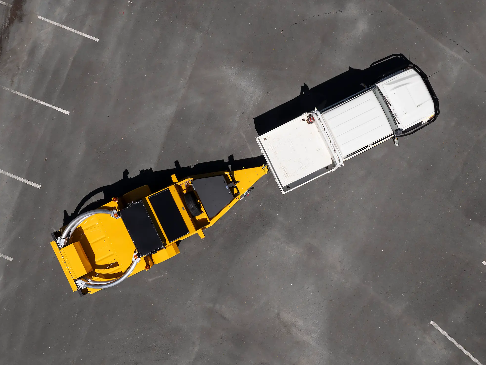 A white utility vehicle is parked in an empty parking lot, towing a large yellow industrial trailer. The image is taken from directly above, casting strong shadows on the pavement.