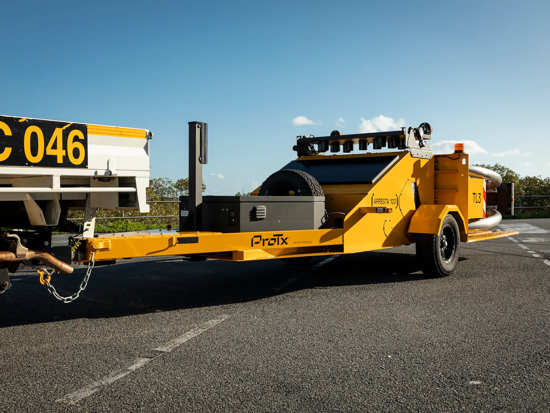 A yellow crash attenuator trailer labeled ProTx is attached to the back of a truck with license plate FC 046, parked on an asphalt road under a clear blue sky.
