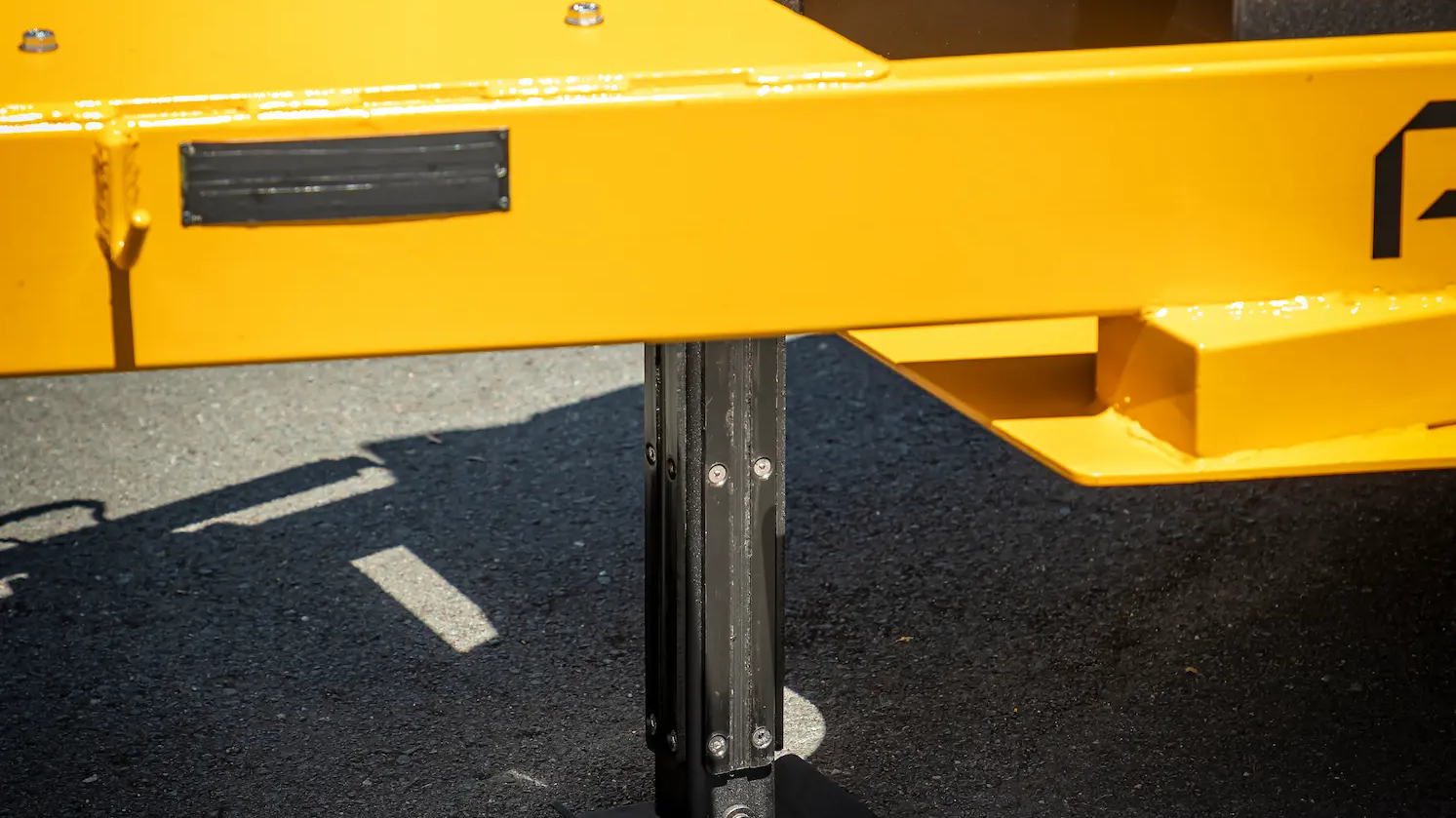 Close-up of a yellow utility trailer’s stabilizer leg extended to the ground, providing support. The trailer is parked on a paved surface, and a black storage box is visible in the background.