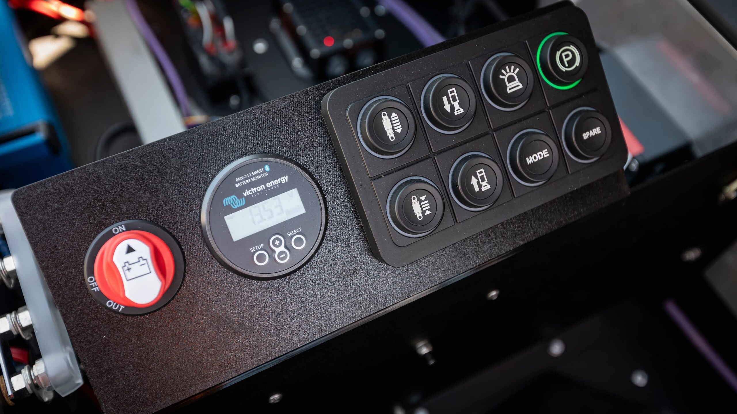 Close-up of an electronic control panel featuring a digital display, a red safety switch, and several labeled black buttons including headlight, horn, and parking symbols, with indicator lights and wiring visible in the background.