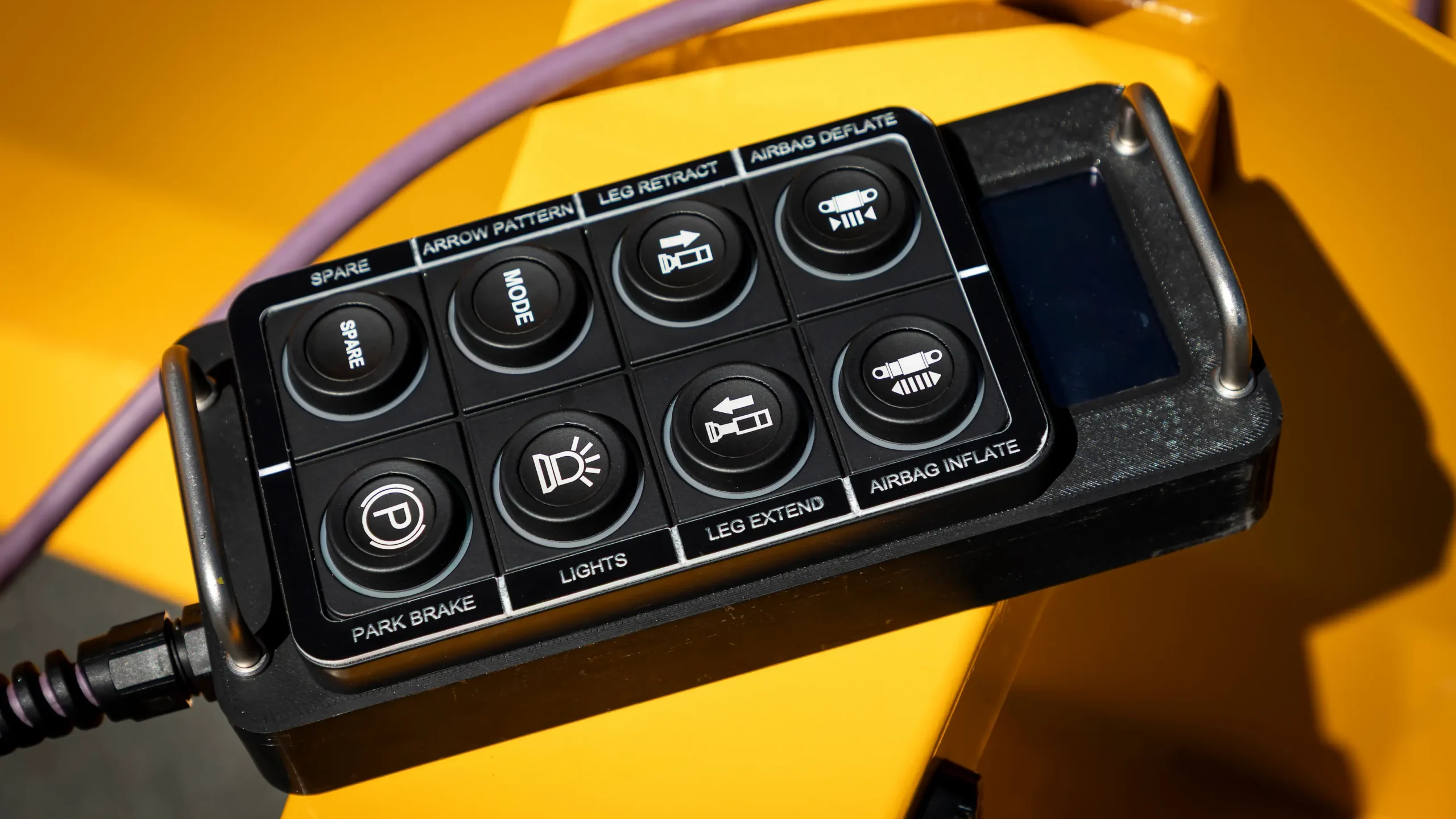 A close-up of a black remote control with labeled buttons, including SPARE, MODE, PARK BRAKE, LIGHTS, ARROW PATTERN, LEG RETRACT, LEG EXTEND, and AIRBAG INFLATE, resting on yellow equipment.
