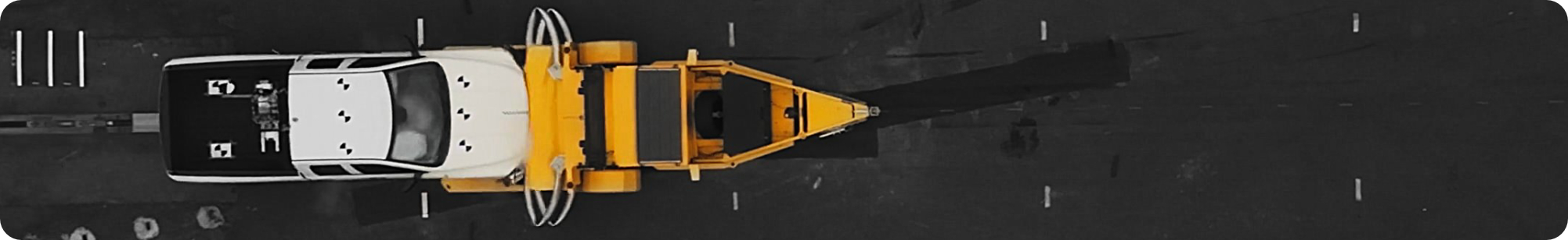 A top-down view of a white vehicle with a yellow tow attachment, positioned on a dark road with white lane markings visible.