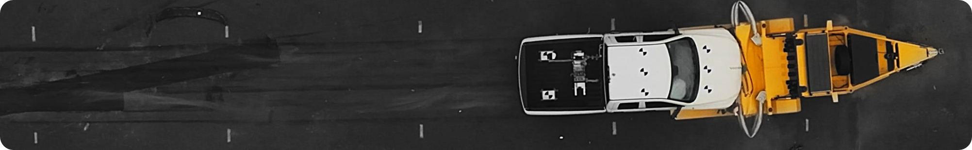 Aerial view of a white vehicle towing a bright yellow crash barrier along a road, with black tire marks visible on the dark pavement.