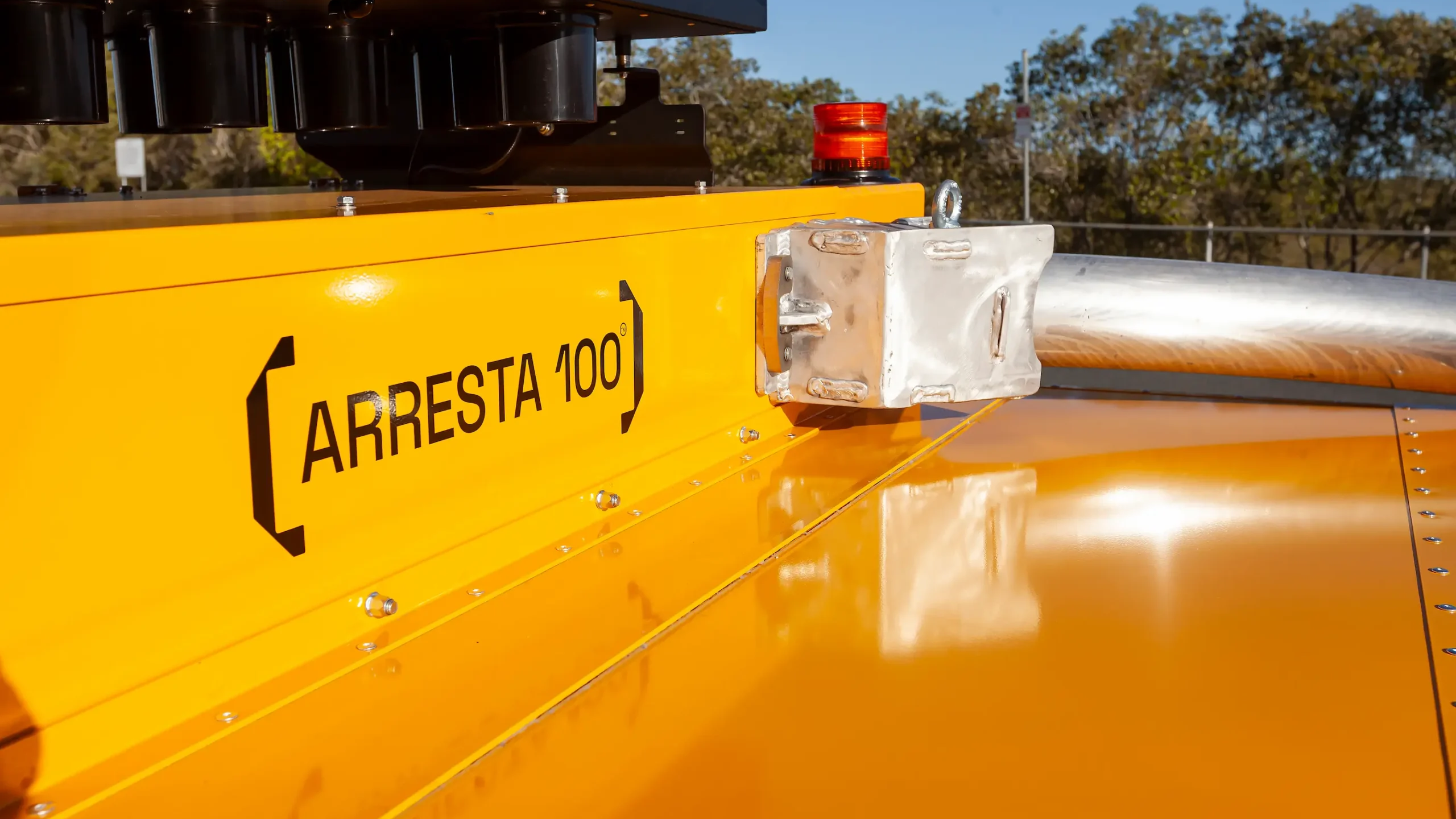 Close-up of a bright yellow vehicle or machine with the label “ARRESTA 100” in black text, a silver metal component, a red warning light, and trees in the background under a clear blue sky.