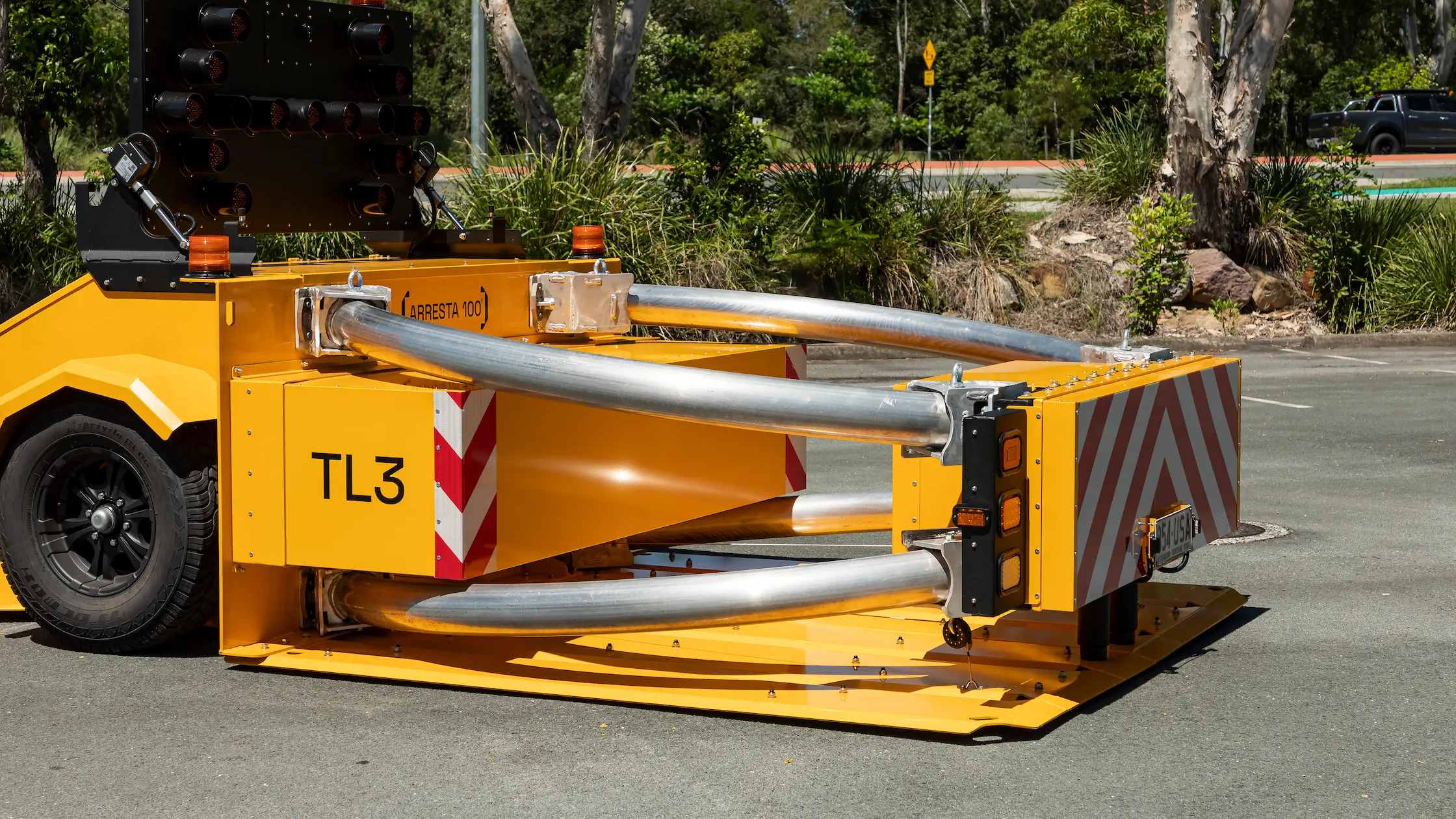 A yellow impact attenuator crash cushion with metal rails and red-white reflective markings is positioned on a paved road, surrounded by greenery and trees.