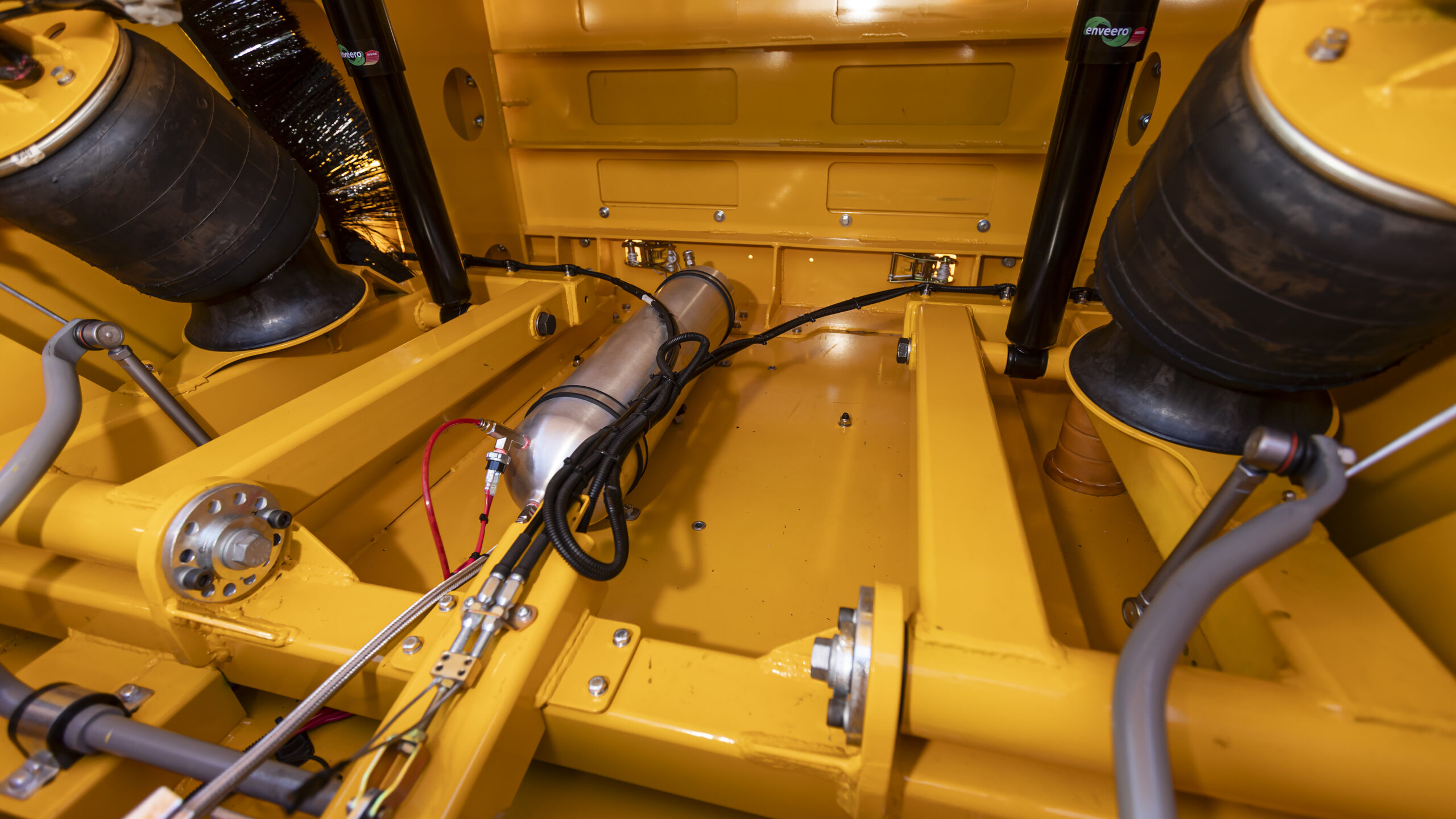 Close-up view of a yellow vehicle’s suspension system featuring large air springs, metal rods, hydraulic components, and wiring inside a metal frame.